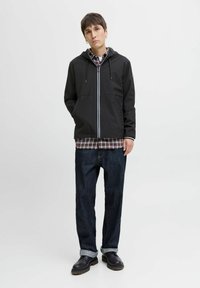 Veste à capuche noire avec des accents blancs, superposée sur une chemise à carreaux, accompagnée de jeans foncés. Le mannequin porte des chaussures noires avec une finition texturée.