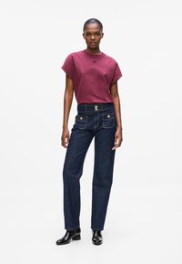 Maroon short-sleeve t-shirt with black logo, paired with high-waisted dark denim jeans featuring two front pockets and gold accents. Black boots.