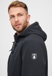 Veste à capuche marine en tissu lisse, dotée d'une fermeture à cordon de serrage et d'un patch logo sur la manche gauche. Doublure contrastée à l'intérieur de la capuche.