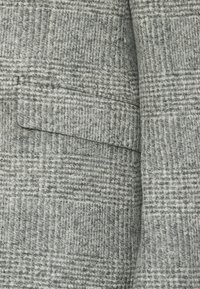 Szary materiał wełniany w mieszance z wzorem w kratkę; posiada zagięty kołnierzyk i boczną kieszeń. Tekstura wydaje się miękka i nieco masywna.