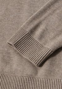Primo piano di una manica e orlo di un maglione lavorato a maglia beige che mostrano i polsini a coste e una texture fine.
