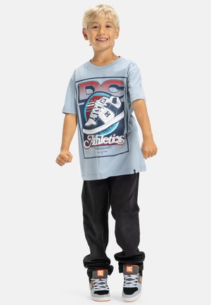 Un garçon souriant portant un t-shirt DC Athletics bleu clair, un pantalon noir et des baskets DC gris-orange, se tenant avec les poings serrés sur un fond blanc.