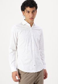 Cămașă albă cu mâneci lungi, cu un model paisley texturat, cu închidere butonată și guler clasic, asortată cu pantaloni de culoare deschisă.