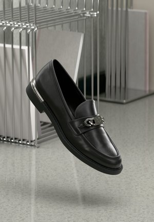 Des mocassins en cuir noir avec un bout arrondi, une semelle plate et un accent métallique sur la sangle. La chaussure présente une texture lisse et une finition polie.