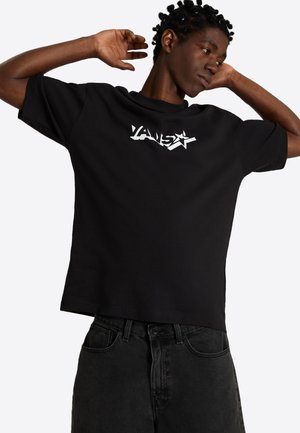 Jeune personne aux cheveux courts torsadés porte un T-shirt Vans noir et un jean foncé, levant les deux bras derrière la tête, regardant sur le côté.