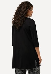 Schwarze Tunika mit Dreiviertelärmeln, seitlichen Schlitzen, glatter Textur und lockerer Passform, kombiniert mit gemusterten Hosen.