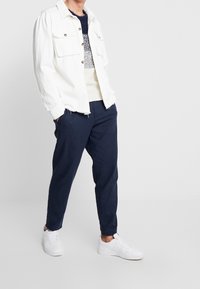 Witte denim jacket met twee voorkruiszakken, gedragen over een textuur van een crèmekleurig en marineblauw sweater, gecombineerd met marineblauwe broek en witte sneakers.
