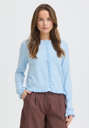 Frau mit langen braunen Haaren, die eine hellblaue, geknöpfte Strickjacke mit Rüschen und braune Hosen trägt, steht mit einer Hand in der Tasche.