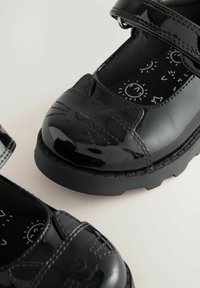 Chaussures en cuir verni noir avec un bout rond, ornées d'un design de visage de chat, avec une sangle réglable et une semelle en caoutchouc noire. Doublure intérieure à motifs.