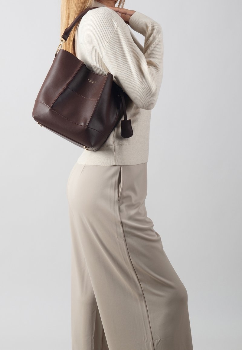 Femme en pull crème et pantalon beige, portant un sac à bandoulière en cuir marron foncé sur l'épaule droite, sur un fond uni.