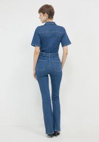 Combinaison en denim à manches courtes, taille ceinturée et jambes évasées. Couleur bleu foncé avec des coutures contrastantes et deux poches arrière.