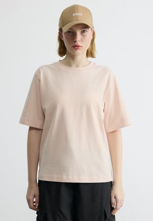 Νεαρό άτομο που φοράει μπεζ καπέλο με τη λέξη "BOSS", ανοιχτό ροζ φαρδιά μπλούζα με μικρό λογότυπο "BOSS" και μαύρο παντελόνι cargo, στέκεται μπροστά σε λευκό φόντο.