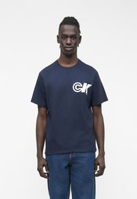 Young man with short dreadlocks wearing a navy blue T-shirt with white "CK" logo and blue jeans against a plain white background.