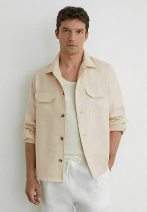 Mand iført beige skjorte med knapper over hvid strikket top og hvide bukser med snøre, stående mod en ensartet lys væg.