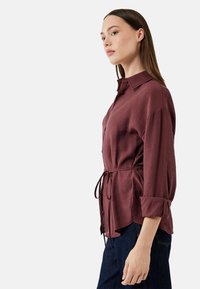 Bordeaux knap-up skjorte med bånd i taljen, blød tekstur, oprullede ærmer og en afslappet pasform, båret over mørk denim jeans.