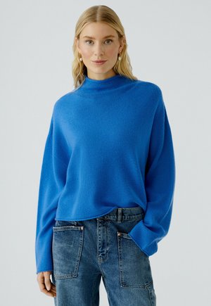 Blauer kuscheliger Pullover mit hohem Kragen und überschnittenen Schultern, kombiniert mit Jeans in entspannter Passform und mit Eingrifftaschen.