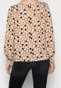 Blouse à manches longues avec un motif géométrique en marron, blanc et noir. Tissu doux, encolure ronde et poignets froncés.