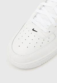 Λευκό δερμάτινο αθλητικό παπούτσι με διατρήσεις στο μπροστινό μέρος, μαύρο λογότυπο κοντά στα κορδόνια και λευκή σόλα με υφή. Διακριτή ραφή.