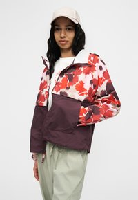 Γυναίκα που φοράει μπουφάν με ροζ και κόκκινο floral μοτίβο, ανοιχτό γκρι παντελόνι με κορδόνι, λευκό πουκάμισο και μπεζ καπέλο μπέιζμπολ, στέκεται μπροστά σε λευκό φόντο.