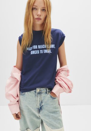 T-shirt en coton bleu marine à manches courtes, avec un texte blanc : "FAIT POUR LES BEACH CLUBS, OBLIGE DE TOUJOURS ENVOYER UN EMAIL." Porté avec un short léger bleu usé.