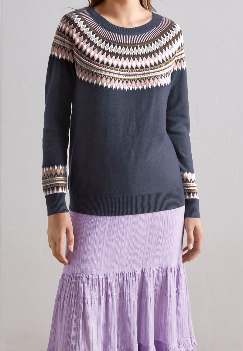 Femme portant un pull bleu marine avec un motif en losanges rose, blanc et marron sur le yoke et les poignets, associé à une jupe à volants violette claire.