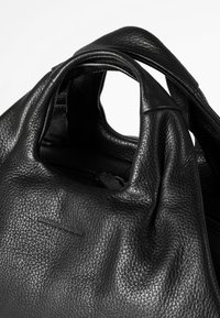 Zwarte leren handtas met een getextureerde afwerking, voorzien van zachte, gedrapeerde hengsels en een minimalistisch logo-detail.
