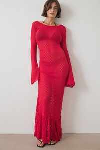 Naine seisab seina vastu, seljas põrandani ulatuv punane võrgukleit pikade varrukatega ja mustad sandaalid, käed lõdvestatult külgedel.