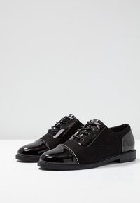 Scarpe nere con tomaia in suede, punta e tallone in vernice, e borchie decorative in argento sul profilo. Dispone di lacci neri e di una suola piatta.