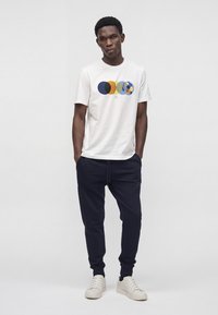 Jeune homme debout, les mains dans les poches d'un jogging bleu marine, portant des baskets blanches et un T-shirt blanc avec un motif de cercles colorés qui se chevauchent.