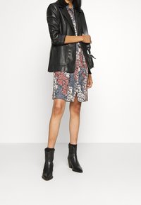 Kvinna klädd i en blommönstrad knälång klänning, svart läderblazer, svarta ankelskor och bärande en svart axelväska mot en vit bakgrund.