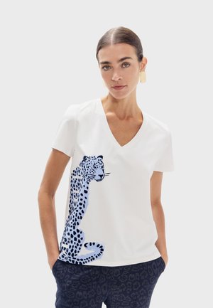 Mujer con camiseta blanca de escote en V con gran estampado de leopardo azul y negro en un lado, combinada con pantalones oscuros con estampado.