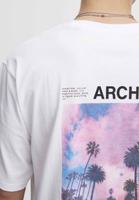 Bagsiden af en hvid skjorte med tekst, delordet "ARCH," en pink og blå himmel samt grafikker af palmer under et sølvfarvet kæde.