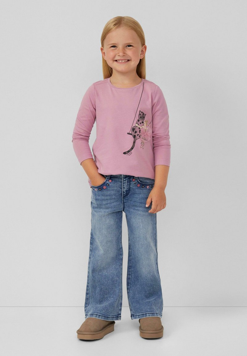Camicia a maniche lunghe rosa chiaro con grafica di un animale, abbinata a jeans svasati blu decorati con accenti floreali. Scarpe marroni completano il look.