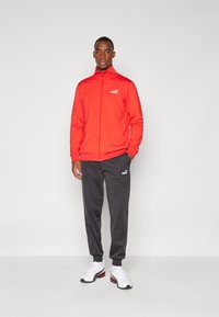 Rode zip-up jack gemaakt van lichtgewicht materiaal, gecombineerd met donkergrijze broek. Bevat Puma-logo op beide items. Sneakers zijn wit met zwarte accenten.