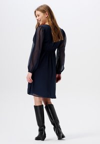 Marineblaues Kleid mit transparenten langen Ärmeln und gerafftem Taillenbereich, kombiniert mit kniehohen schwarzen Lederstiefeln, die eine glatte Textur und modernes Design zeigen.