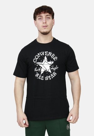 Giovane uomo che indossa una t-shirt nera Converse All Star con logo a stella frammentato e pantaloni verde scuro su sfondo bianco uniforme.