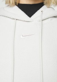 Vit fleecehoodie med en strukturerad yta, justerbar dragsko huva och en liten silverfärgad Nike-logotyp på bröstet.