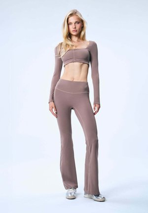 Giovane donna in piedi che indossa un crop top a maniche lunghe color malva e pantaloni a zampa abbinati con sneakers argento su uno sfondo chiaro e uniforme.