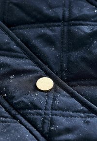 Tessuto trapuntato blu navy con gocce d'acqua, caratterizzato da un grande bottone in metallo dorato e motivi di cuciture diagonali. Texture liscia, materiale resistente.