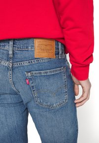 Jean bleu en denim avec une pièce en cuir portant l'inscription "Levi Strauss & Co." et un petit onglet rouge sur la poche arrière, associé à une chemise rouge à manches longues.