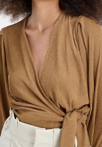 Top avvolgente marrone a coste con maniche lunghe e una cintura in vita, abbinato a una cintura bianca. Il tessuto testurizzato crea un aspetto morbido e stratificato.