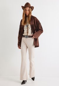 Bruin suède jack met franjes, crèmekleurig graphic t-shirt, beige flare broek en zwarte puntige hakken; accessoires zijn een bruine hoed en een zilveren riem.