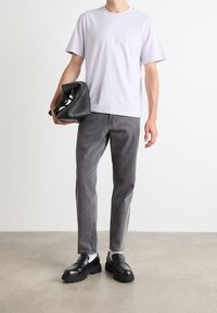 Osoba ubrana w jasnofioletowy T-shirt, szare dżinsy, czarne mokasyny z białymi skarpetkami, trzymająca złożoną czarną kurtkę w lewej ręce.