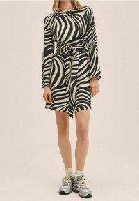 Femme portant une robe à manches longues à rayures zèbre noir et blanc avec une ceinture nouée à la taille, des chaussettes blanches et des baskets de sport grises, debout devant un fond uni.