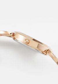 Orologio da polso in oro rosa con cassa rotonda, corona testurizzata e finitura lucida; presenta un cinturino sottile e un vetro trasparente.