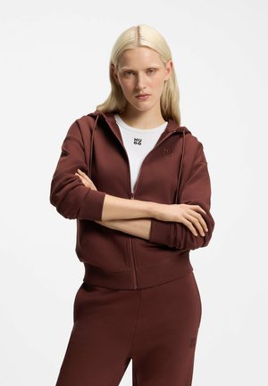 Lyshåret kvinde med armene over kors iført en brun lynlåshoodie og matchende bukser over en hvid skjorte med Hugo-logo på brystet.