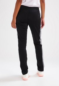 Svarta träningsbyxor med elastisk midja, med vita ränder längs sidorna. Materialet verkar smidigt och något reflekterande.