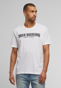Bílé bavlněné tričko s krátkým rukávem, na kterém je výrazný černý nápis "TRUE RELIGION BRAND JEANS" přes hruď.
