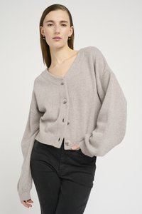Cardigan corto grigio chiaro con scollatura a V profonda, maniche oversize e bottoni grandi. Il tessuto sembra morbido e strutturato. Abbinato a pantaloni neri.