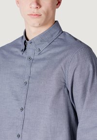 Camicia da uomo con bottoni in tessuto azzurro chiaro struttura, con colletto classico e bottoni scuri. Stemma ricamato discreto vicino al petto.